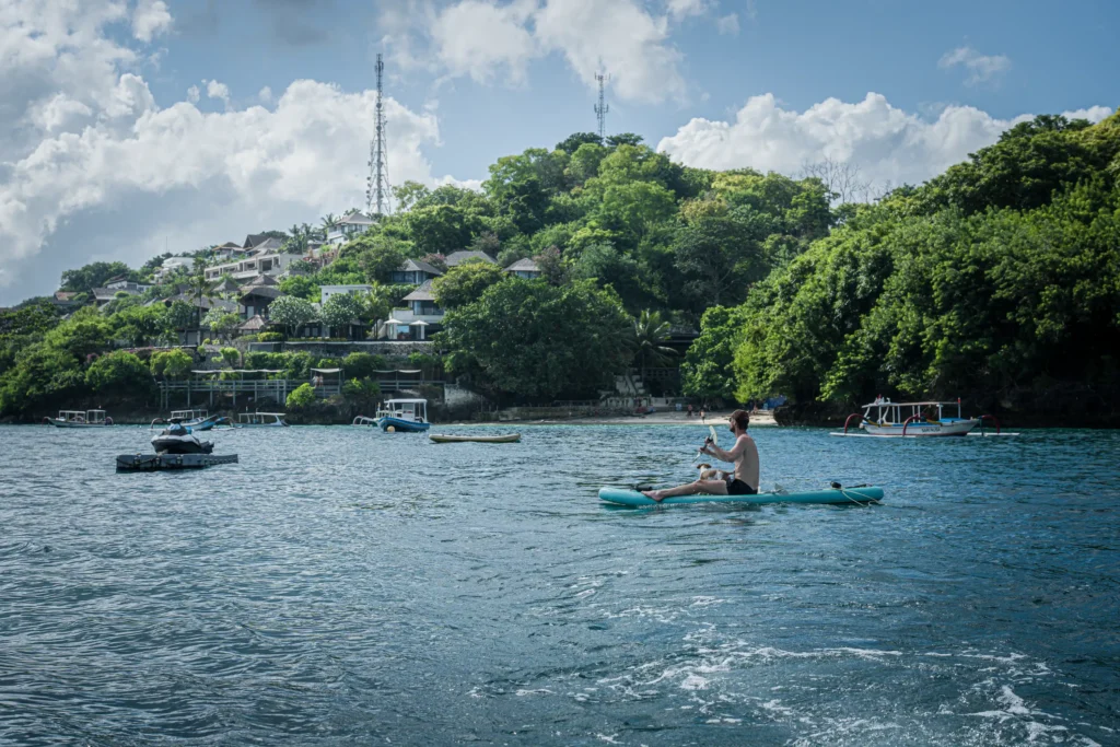 A guest paddles a clear kayak across calm water off Mushroom Bay, Nusa Lembongan, with traditional jukung boats and a hillside of cliffside villas behind.