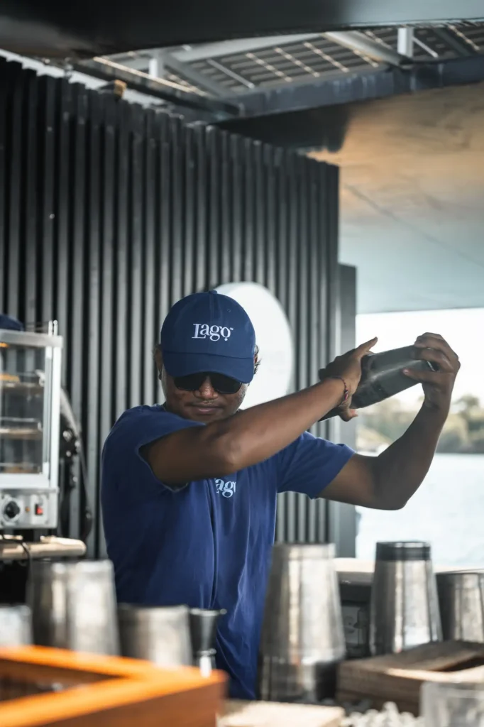 A Lago bartender in branded navy cap and tee shakes a cocktail behind the bar at the Pontoon at Lago.