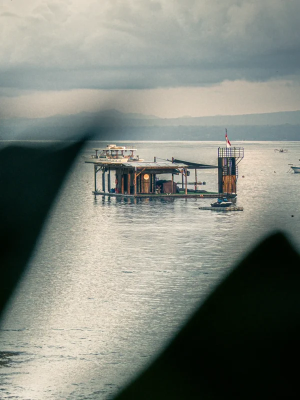 The Pontoon at Lago floating bar seen from a distance across the bay, framed by a traditional sail under moody overcast skies.