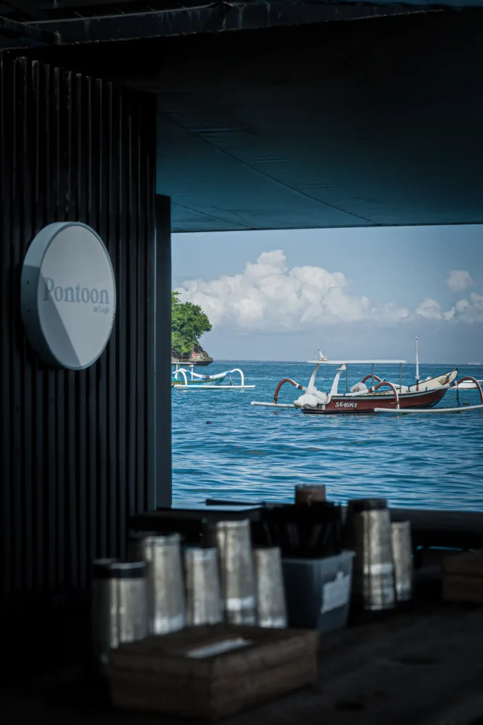 The Pontoon at Lago circular signage on dark timber walls, looking out to traditional Balinese jukung outrigger fishing boats moored on the bay.