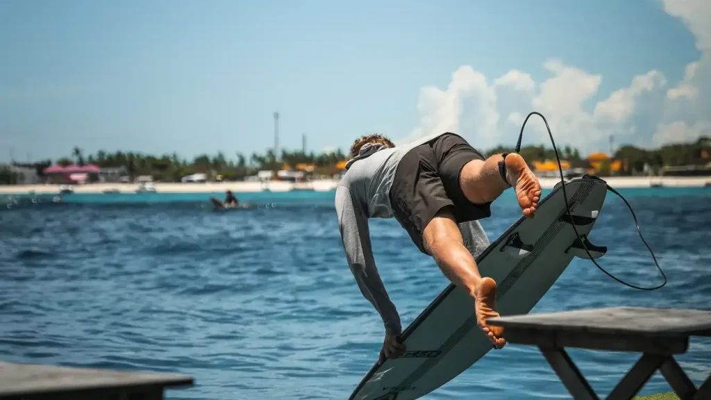 A surfer leaps from the astroturf deck of the Pontoon at Lago with board in hand, crystal-clear water below and the palm-fringed Lembongan shore beyond.
