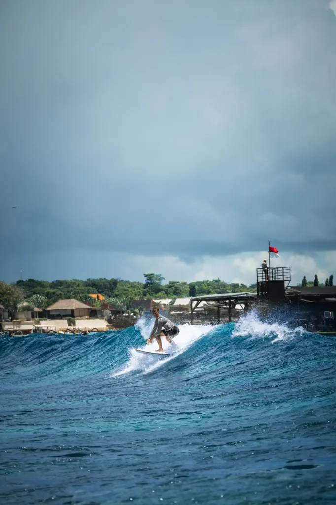 A surfer rides a clean blue wave with the Pontoon at Lago floating bar and Indonesian flag silhouetted against moody skies in the background.
